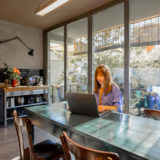 A woman in a relaxed blue outfit works on her laptop at a rustic dining table in a modern kitchen. Large glass windows fill the space with natural light, creating a serene and productive atmosphere